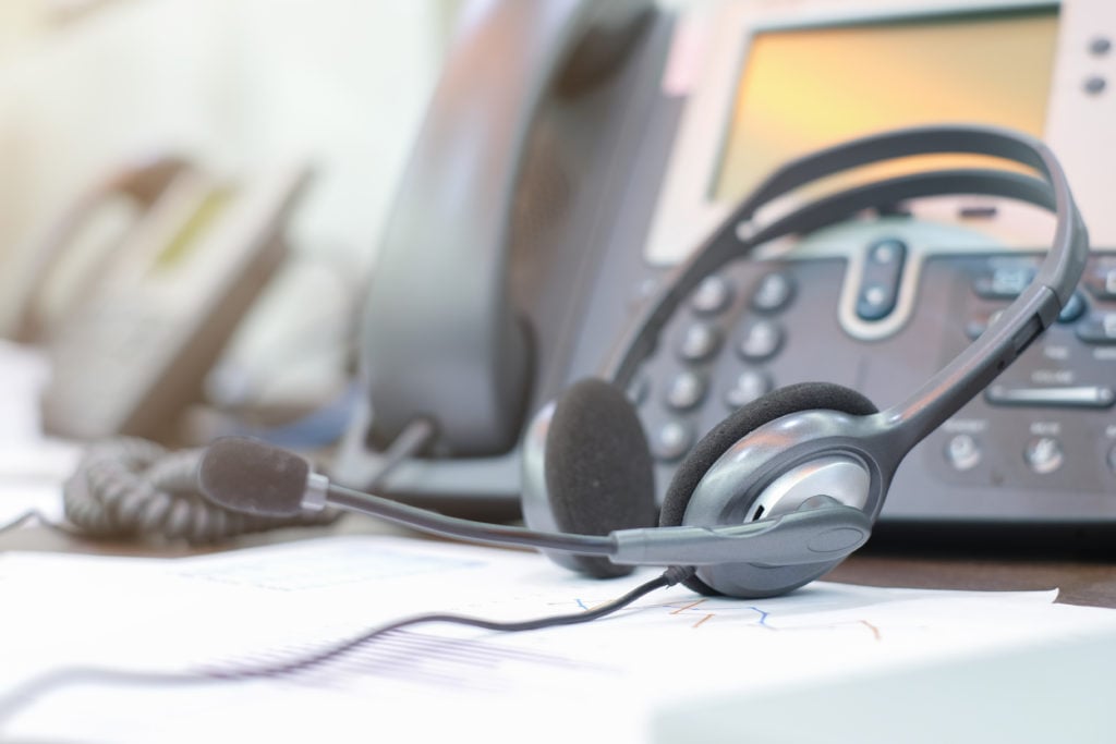 A headset with a microphone rests on a desk next to an office phone, hinting at the seamless communication offered by hosted PBX systems, with another phone and paperwork visible in the background.
