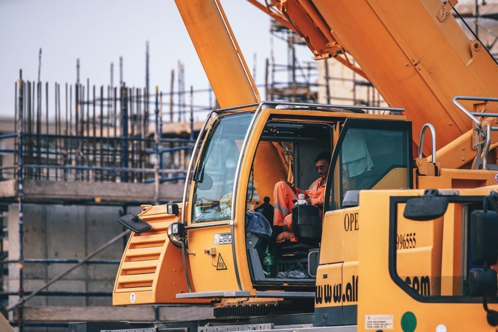 A construction worker in an orange uniform sits inside the cab of a yellow crane, surrounded by scaffolding and building materials, while fleet management software for construction equipment helps streamline operations at the site.
