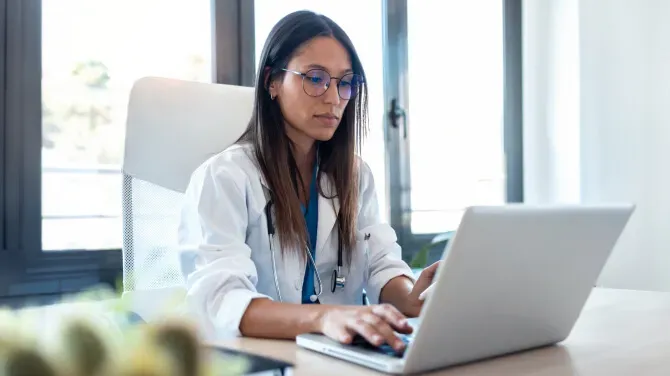 A woman wearing a white lab coat and glasses sits at a desk, typing on a laptop. She appears focused, and a stethoscope hangs around her neck. Large windows are in the background.