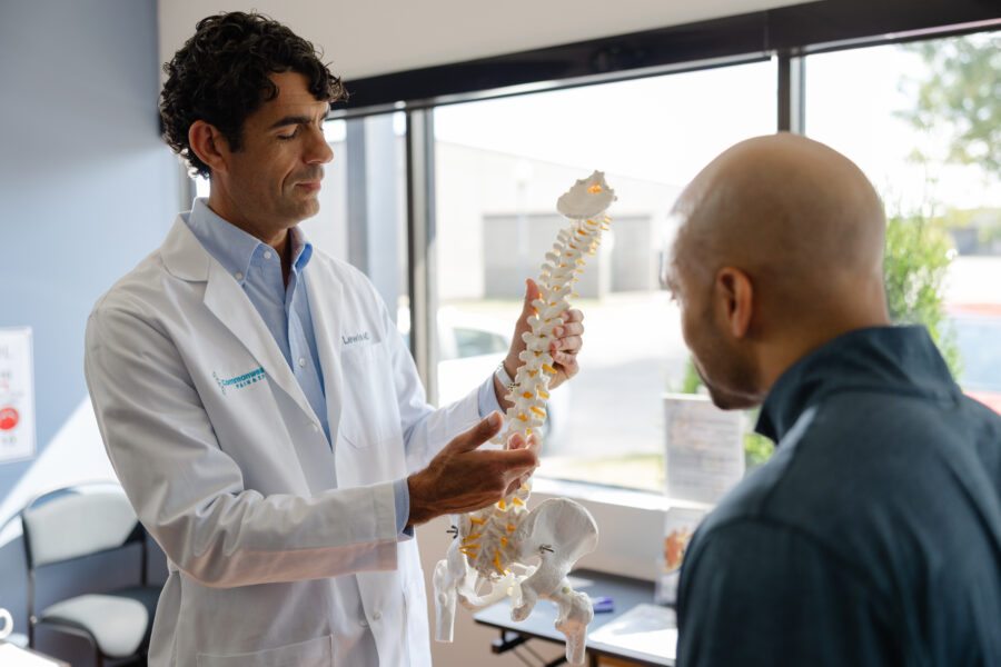 A doctor in a white coat holds and explains a model of a human spine to a patient in a medical office with large windows.