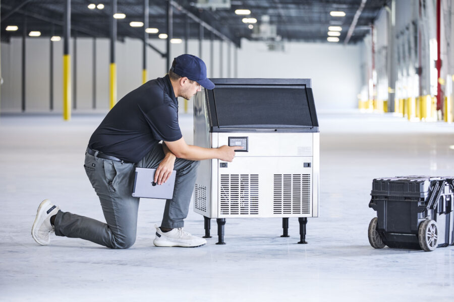 A technician wearing a cap and holding a tablet kneels on the floor in a large warehouse, pressing buttons on a commercial ice machine. A black tool case sits nearby.
