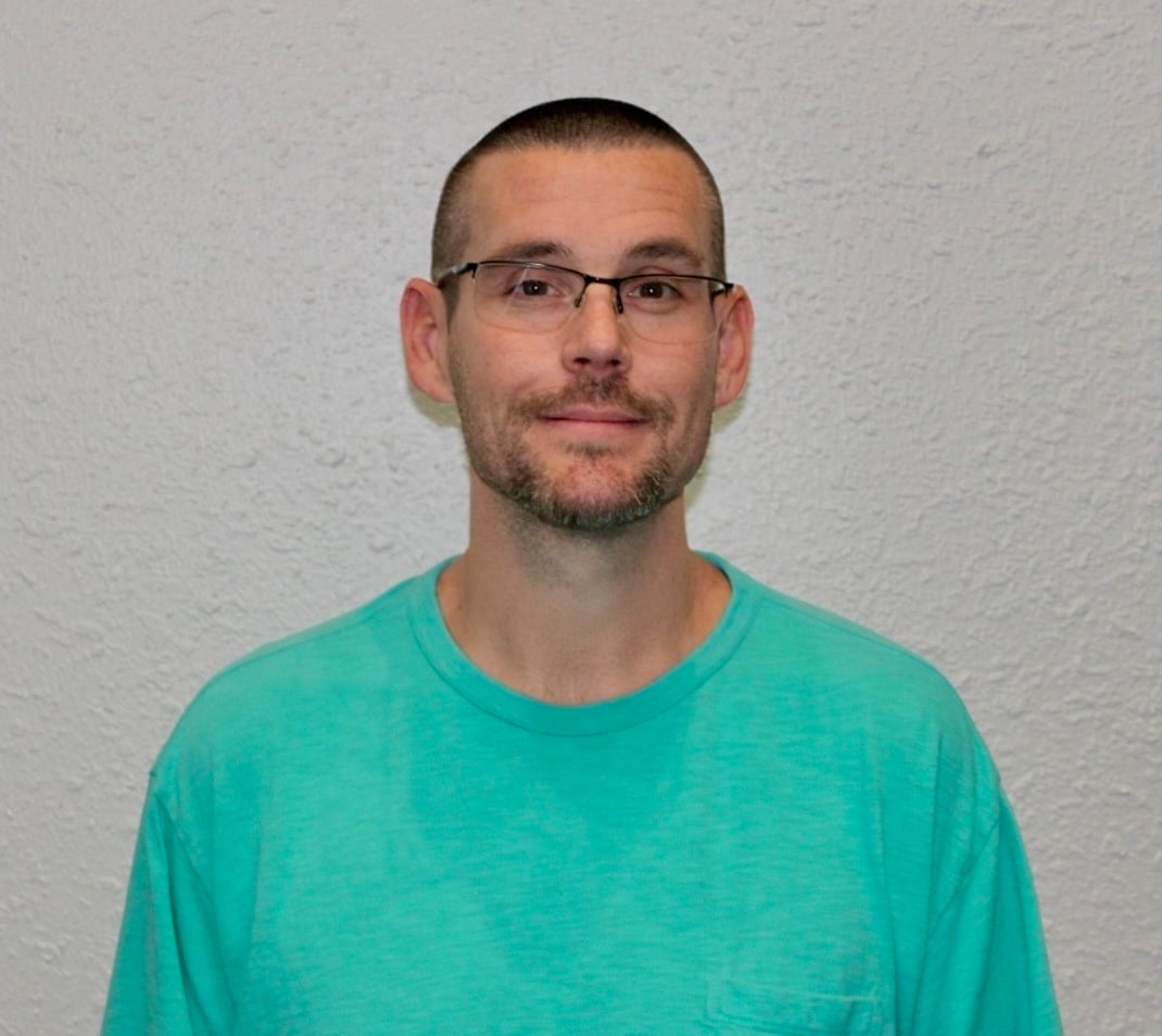 A man with short hair, glasses, and a trimmed beard is standing against a textured white wall, wearing a teal T-shirt and looking at the camera with a neutral expression.