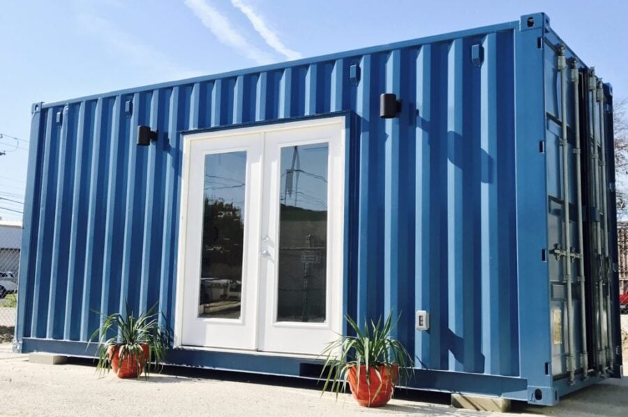 A blue shipping container converted into a small building with white double doors and two potted plants in red pots placed on either side of the entrance.