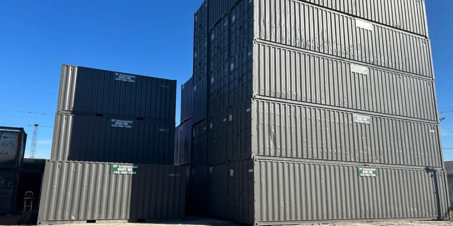 Tall stacks of large gray shipping containers are piled outdoors under a clear blue sky. The containers are arranged in various heights, creating a geometric shape in the open area.