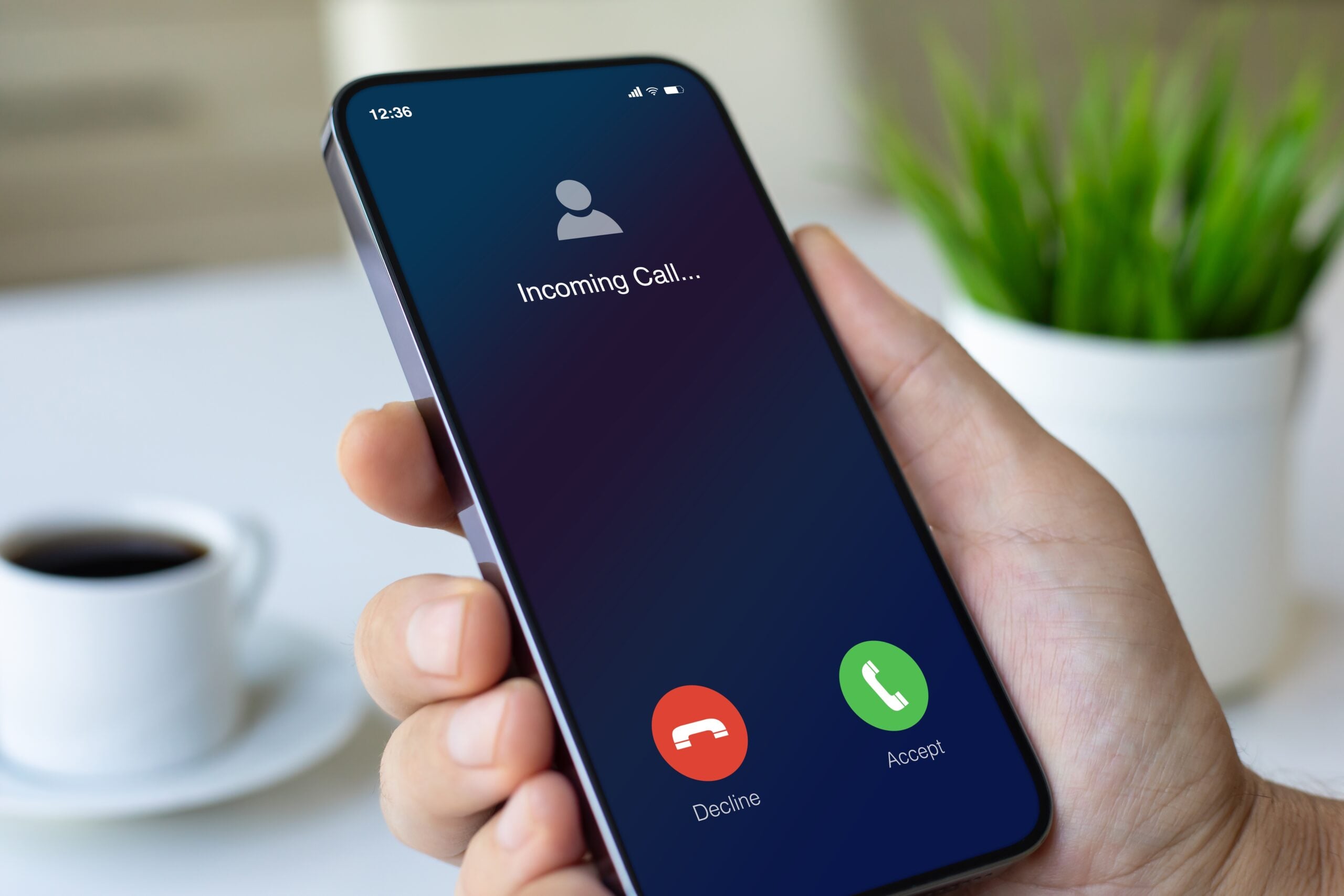 A hand holds a smartphone displaying an incoming call screen with Decline and Accept buttons. A cup of coffee and a green plant are blurred in the background.