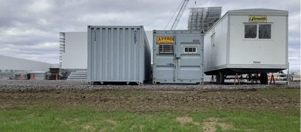 Three portable structures sit on gravel at a construction site: two shipping containers and one white trailer, with construction materials and a crane in the background under a cloudy sky.