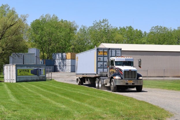 A large truck is transporting a shipping container on a gravel road beside stacked containers and a warehouse, with green grass and trees in the background.