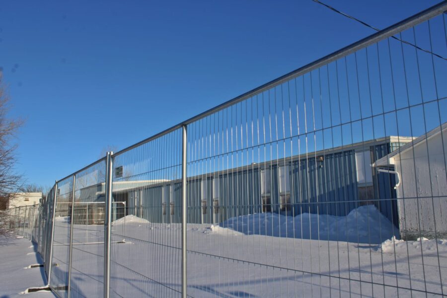 A metal fence surrounds a blue industrial building with snow on the ground under a clear blue sky.