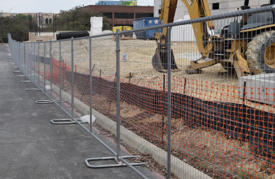 Construction site with a metal fence, orange safety netting, dirt, large machinery, and building materials. A street runs alongside the fenced-off area. Buildings and containers are visible in the background.