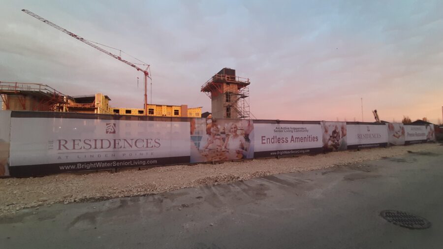 A construction site at sunset, with cranes and unfinished buildings in the background. Signs on the fence advertise The Residences at Linden Pointe senior living with amenities.