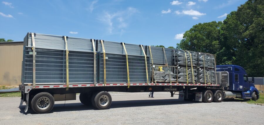 A flatbed trailer loaded with stacked metal fencing materials is secured with yellow straps, parked on gravel near a blue semi-truck under a clear blue sky.