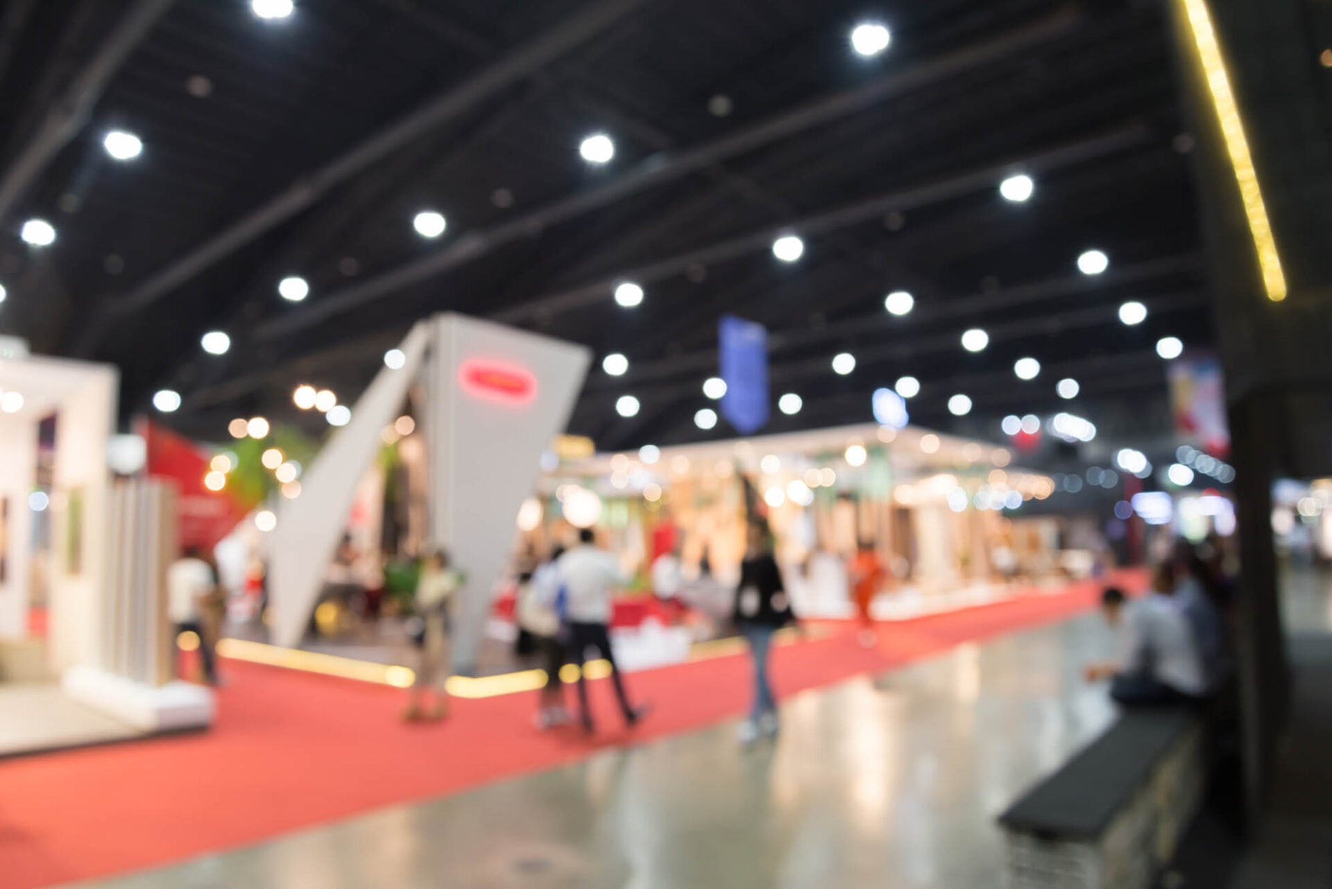 Blurred view of people walking through a large exhibition hall with booths and red carpeting.